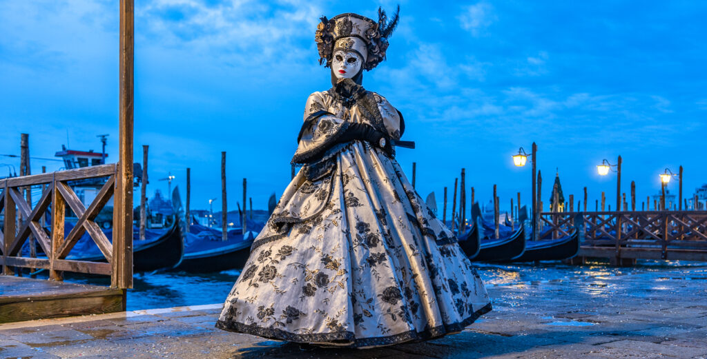 Karneval in Venedig, Fotografie von Christine Hübner-Berlepsch, Frau in traditionellem Kostüm posiert am Ufer vor venezianischen Gondeln, mystische Stimmung durch Morgendämmerung und Blaue Stunde, Laternenlicht erhellt Frau im Kostüm